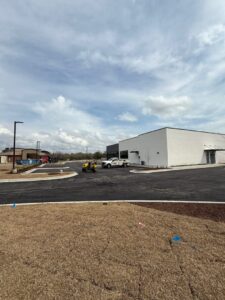 Newly paved commercial parking lot with clean asphalt surface in front of a modern white building, featuring work trucks and paving equipment under a partly cloudy sky.