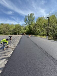 Crew performing asphalt paving on a commercial parking lot surrounded by trees under a clear blue sky. Workers are seen raking and smoothing fresh blacktop pavement during a sunny day.