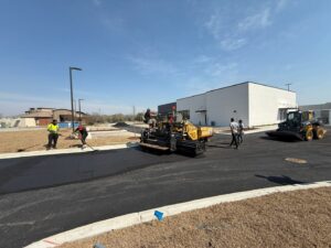 Asphalt paving crew working on a new commercial parking lot. Workers are raking fresh asphalt while a paver machine lays a new surface near a white commercial building under clear blue skies.