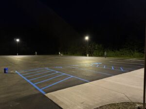 Freshly paved parking lot at night with bright blue ADA-compliant line striping, illuminated by overhead lights as workers finish detailing the site against a dark wooded background.