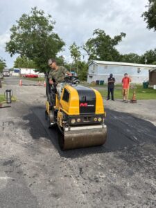 Worker operating a yellow asphalt roller during asphalt repair work in a residential neighborhood, compacting freshly patched pavement with team members assisting in the background.