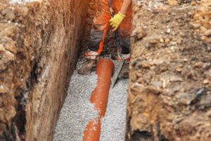 Worker installing a perforated drainage pipe in a gravel-filled trench, preparing proper subsurface drainage to prevent water buildup beneath pavement and asphalt surfaces.