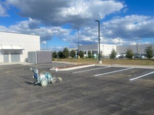 Freshly paved commercial parking lot with newly painted white parking lines and a line striping machine on the asphalt, set beside a modern industrial building under a partly cloudy sky.