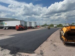 Fresh asphalt being laid and compacted in an industrial lot, with paving equipment and trucks visible under a cloudy sky.