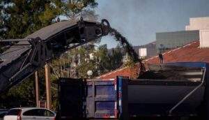 Asphalt milling machine conveyor loading removed pavement into a dump truck during a road resurfacing project.